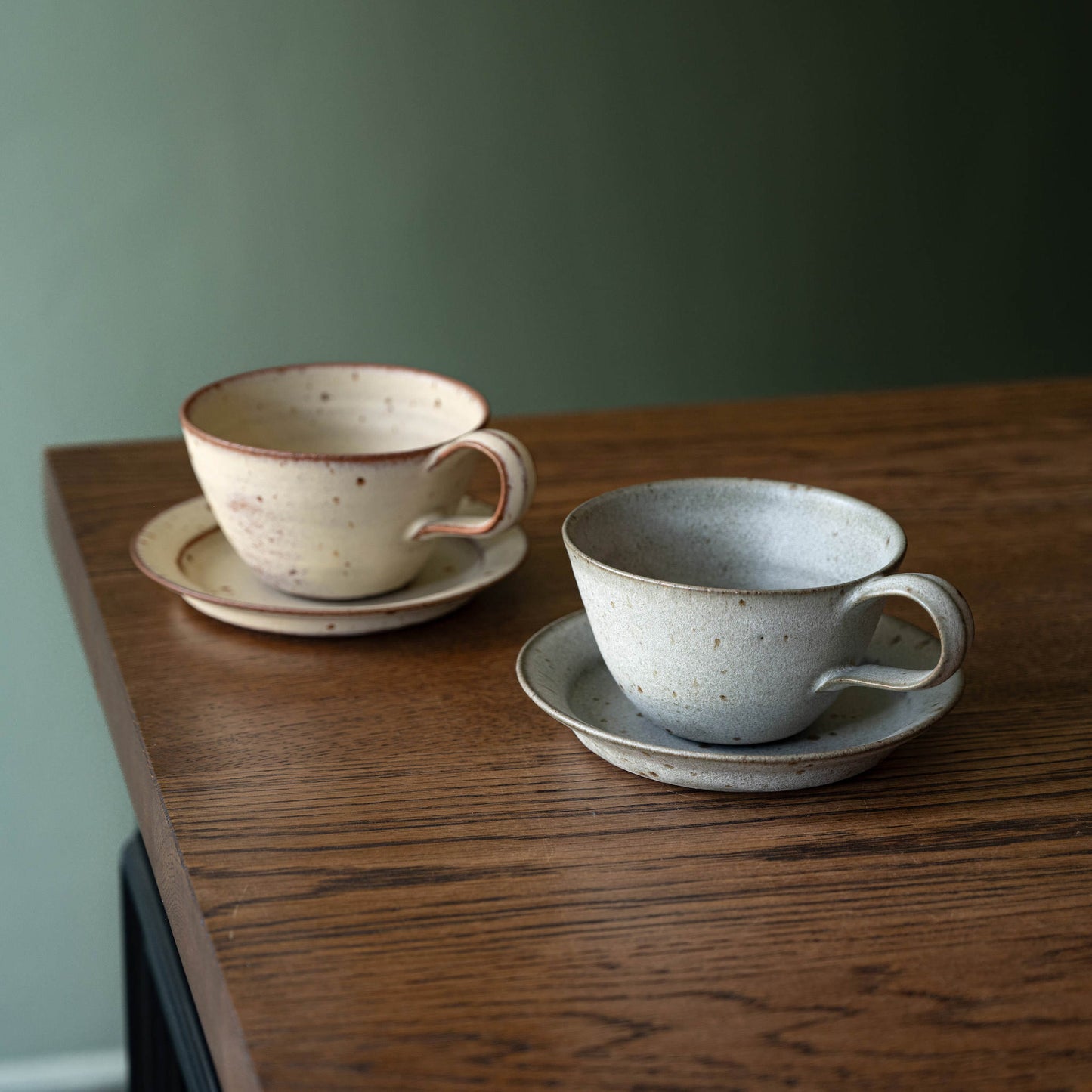 Two handmade ceramic tea cups with saucers / biscuit plate by Studio Brae. One cup is cream-coloured with brown speckles and a reddish-brown rim, while the other is light gray with similar speckles and a brown rim.