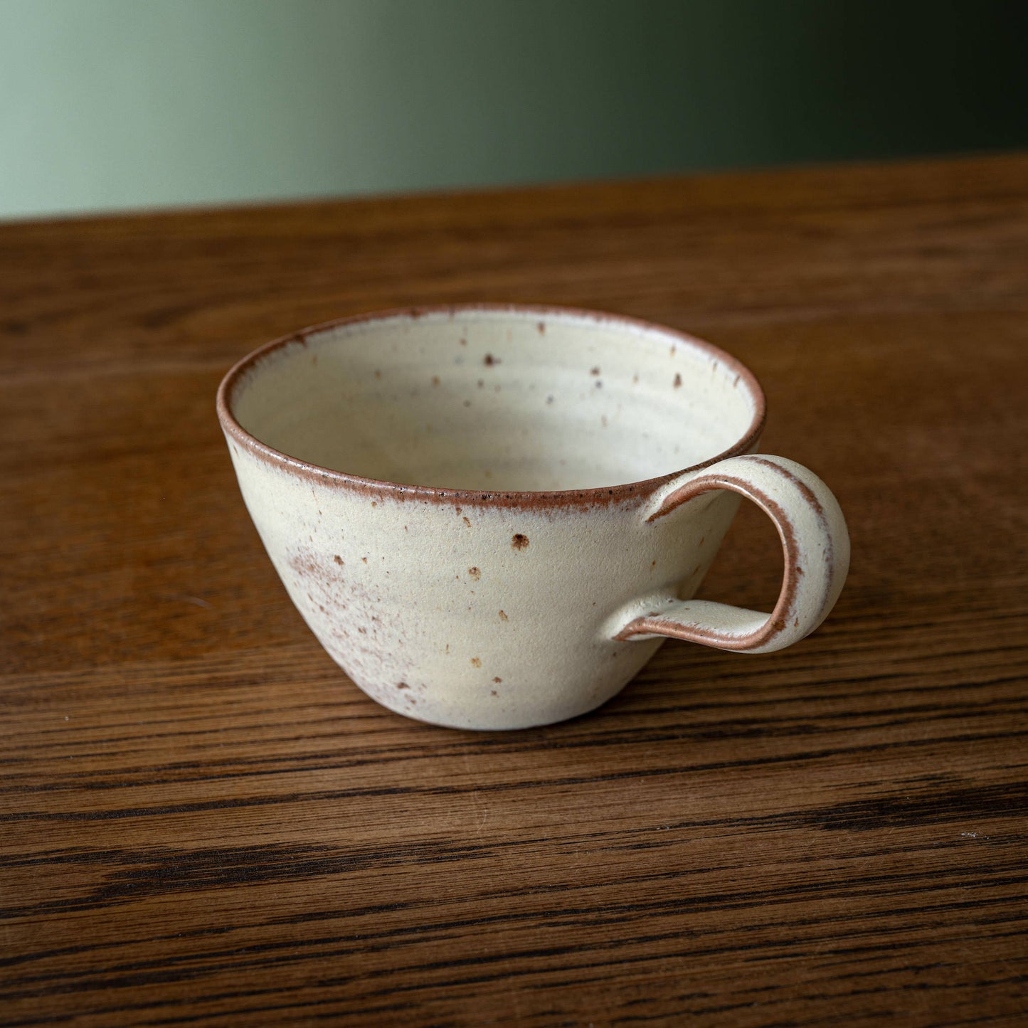 A handmade ceramic curved cup by Studio Brae with a speckled off-white glaze and a rustic reddish-brown rim, shown on a dark wood tabletop.