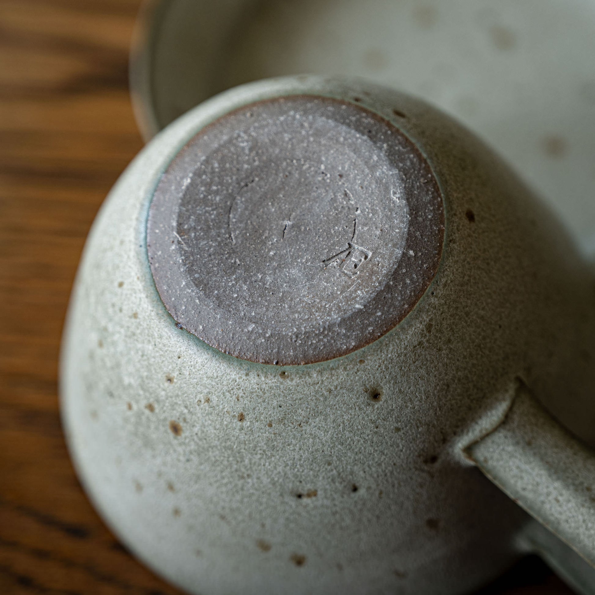 Close-up of the bottom of a speckled ceramic cup, showing a matte gray-brown glazed base, resting on a wooden surface.