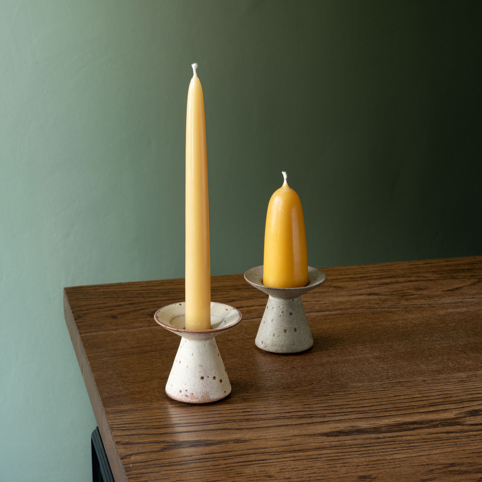 Beeswax dinner candle and beeswax stumpie candle in speckled glaze handmade ceramic holders, displayed on a wooden table against a muted green wall by Studio Brae.