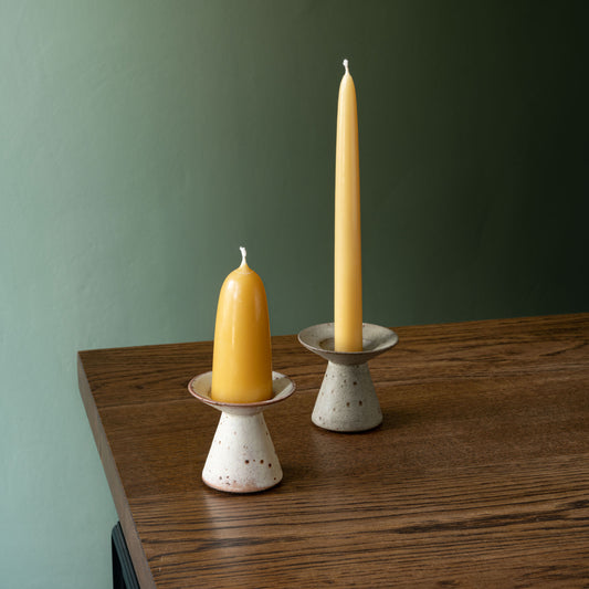 Two handmade ceramic candle holders by Studio Brae on a wooden table against a muted green wall, holding beeswax dinner and conical candles.