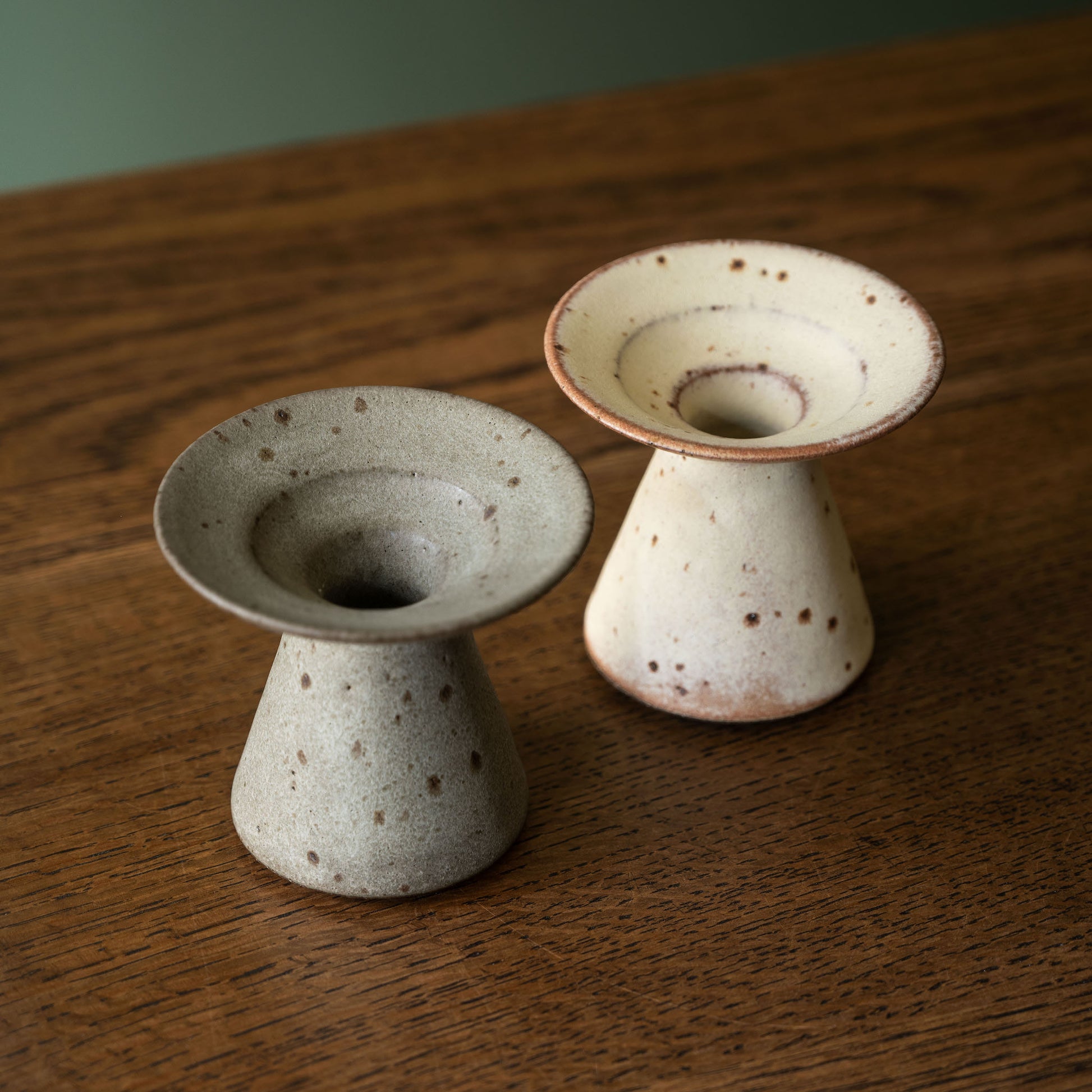 Two handmade ceramic candle holders by Studio Brae with a speckled glaze, one in light grey and the other in cream with brown speckles, displayed on a wooden table.