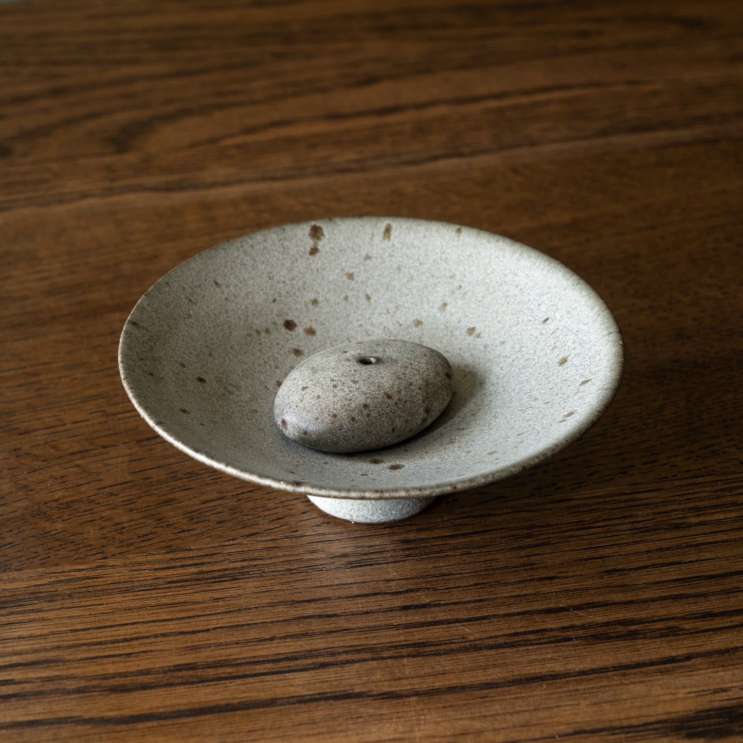 Handmade ceramic incense holder by Studio Brae. A small round ceramic dish with a pedestal base and a smooth grey stone placed in the centre with hole for the incense, set on a dark wood tabletop.