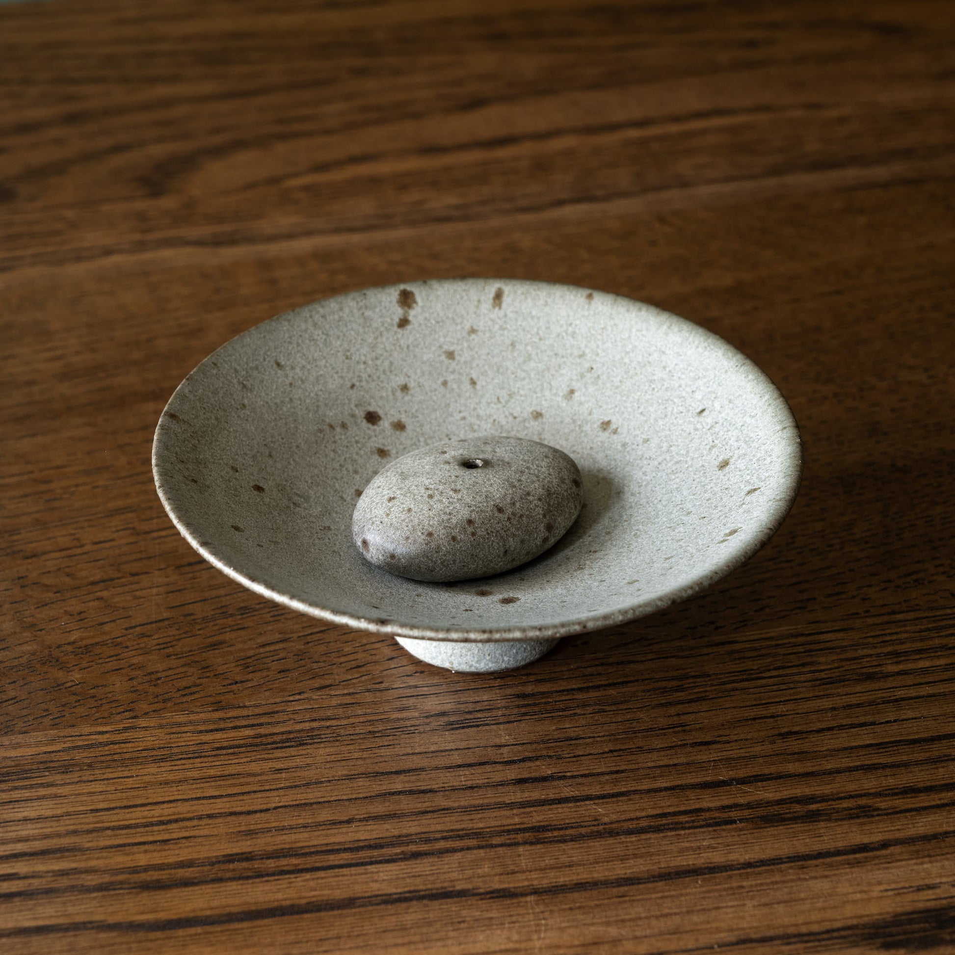 Handmade ceramic incense holder by Studio Brae. A small round ceramic dish with a pedestal base and a smooth grey stone placed in the centre with hole for the incense, set on a dark wood tabletop.