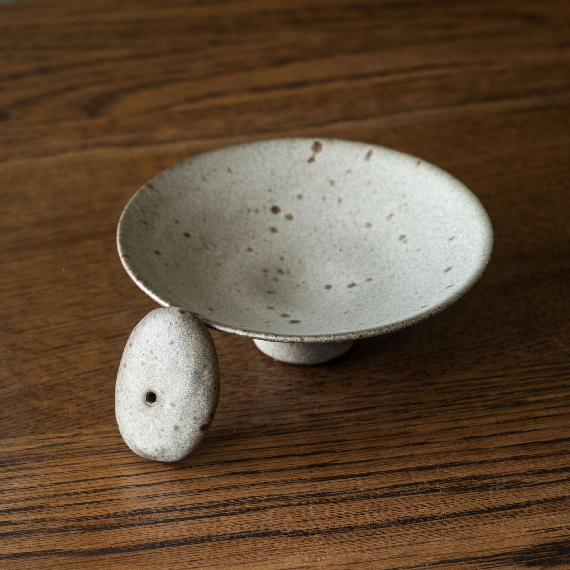 A handmade ceramic footed incense bowl by Studio Brae with a small matching stone pebble incense holder on its side, displayed on a dark wood surface.