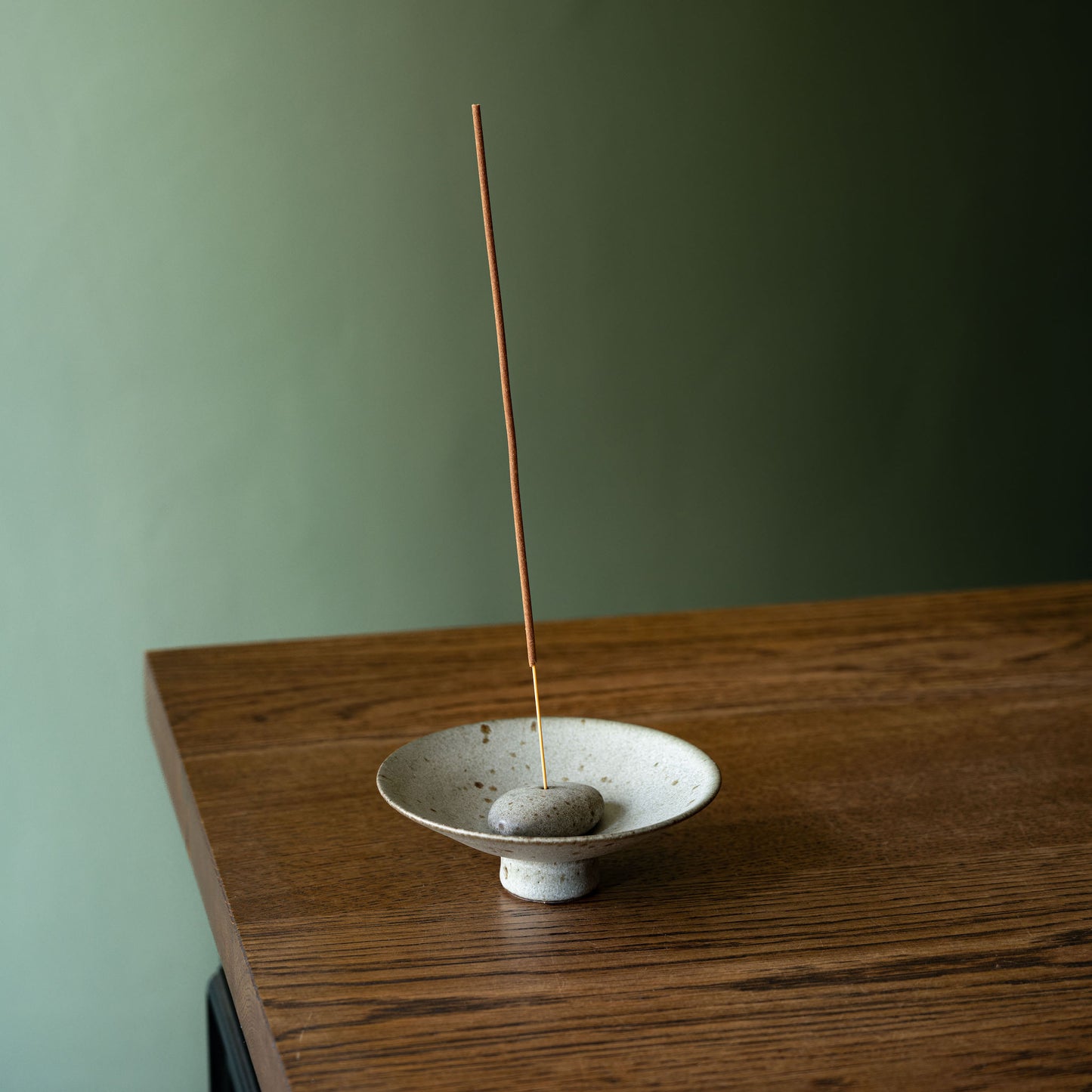 A handmade ceramic incense holder by Studio Brae with a small grey speckled bowl and pebble base, and a single incense stick standing vertically, placed on a wooden table against a muted green wall.