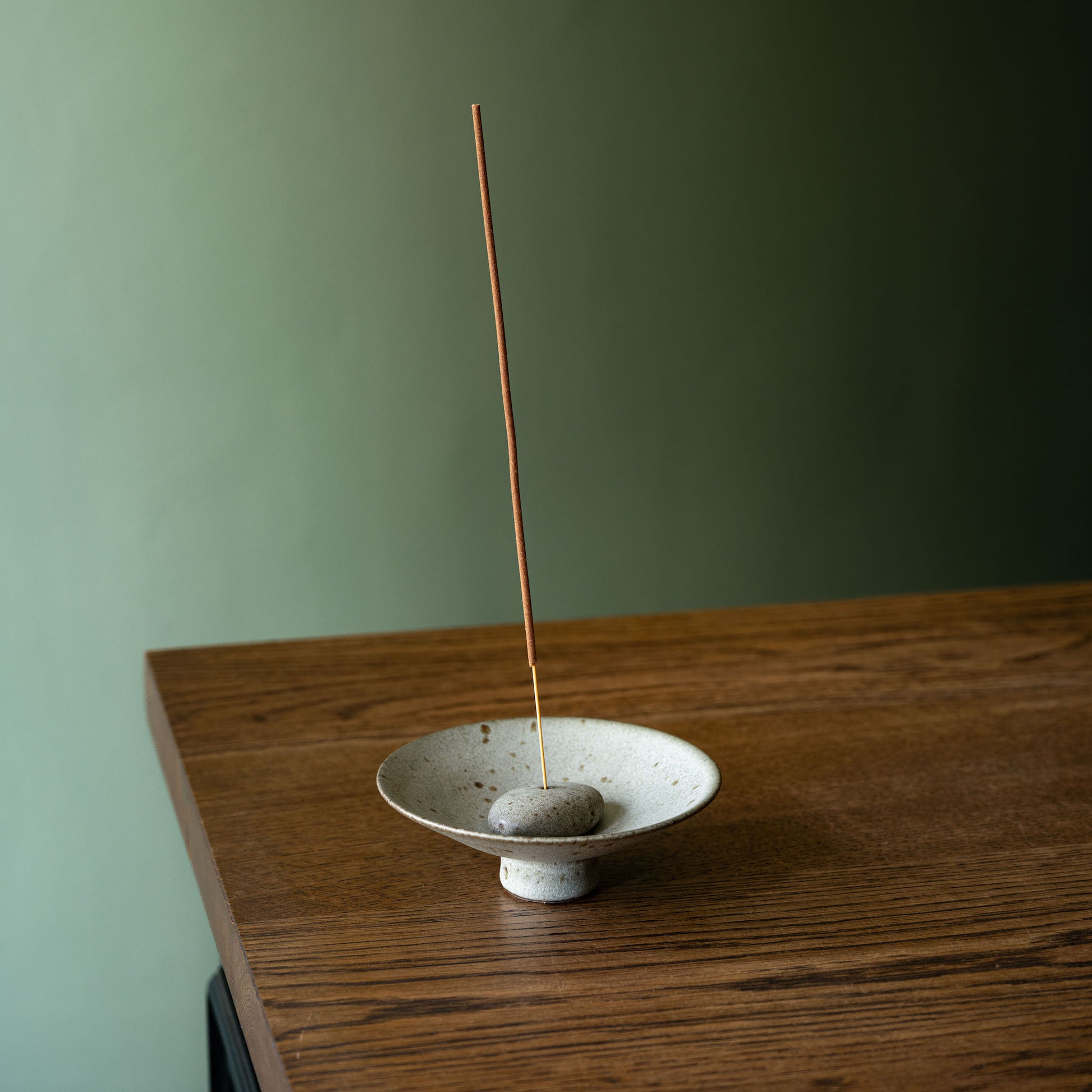 A handmade ceramic incense holder by Studio Brae with a small grey speckled bowl and pebble base, and a single incense stick standing vertically, placed on a wooden table against a muted green wall.