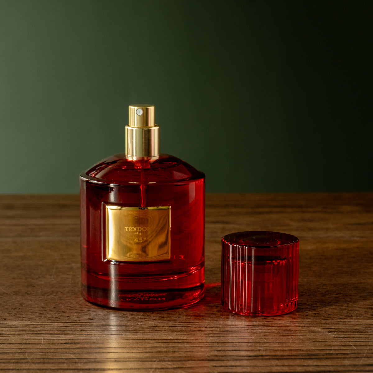 Trudon 45 Eau de Parfum in a red glass bottle with a gold spray top and a matching red ribbed cap, set on a wooden surface against a dark green background. 