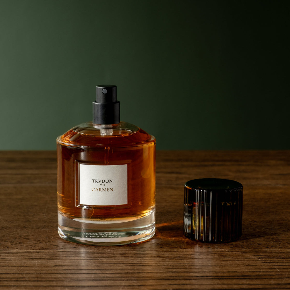 Trudon Carmen Eau de Parfum in a clear glass bottle with a black spray cap, sits on a wooden surface against a dark green background, with its black ribbed cap placed beside it.