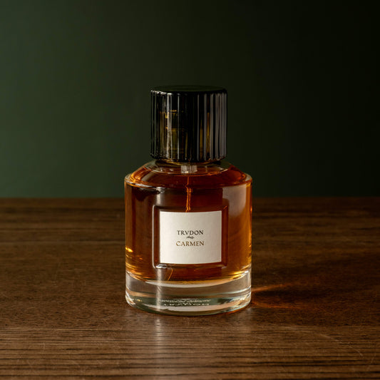 Trudon Carmen Eau de Parfum in a clear glass bottle with a black ribbed cap, standing on a wooden surface against a dark green background. 