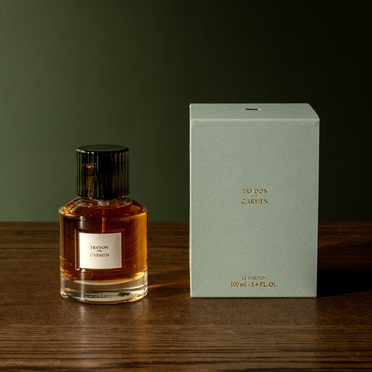 A bottle of Trudon Carmen Eau de Parfum next to its pale blue-green box on a dark wooden surface against a dark olive background.