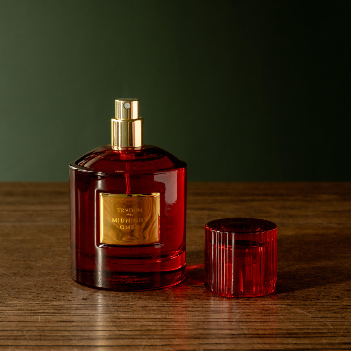 A bottle of Trudon Midnight Omen Eau de Parfum with a gold spray top and a matching red ribbed red cap, set on a wooden surface against a dark green background.