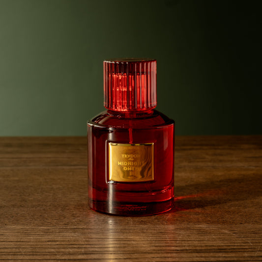 Trudon Midnight Omen Eau de Parfum. A red translucent glass bottle and red ribbed cap, with a gold label reading 'TRUDON MIDNIGHT OMEN' sits on a dark wooden table against a dark green background.