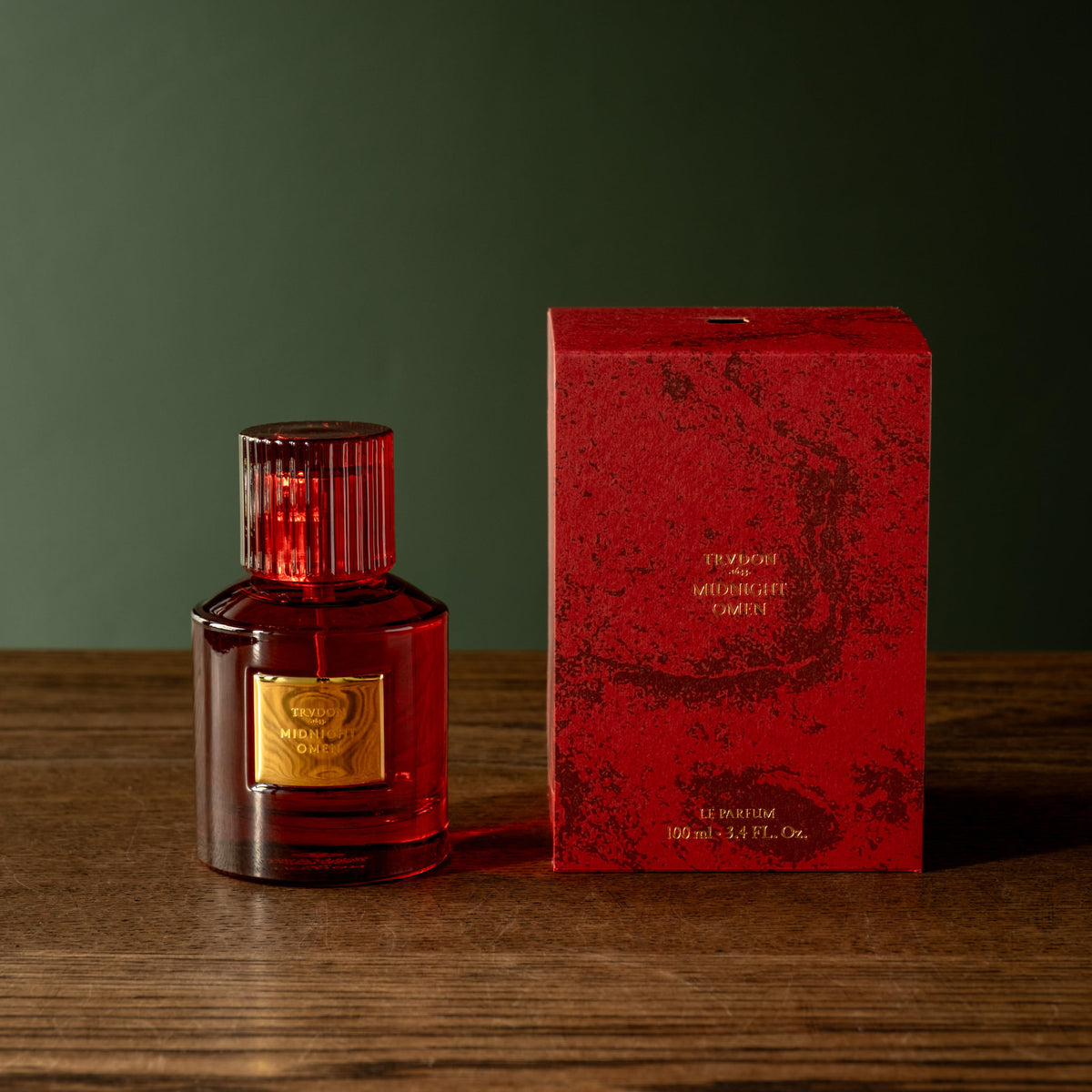 Trudon Midnight Omen Eau de Parfum  stands beside a matching red box on a wooden surface against a dark green background.