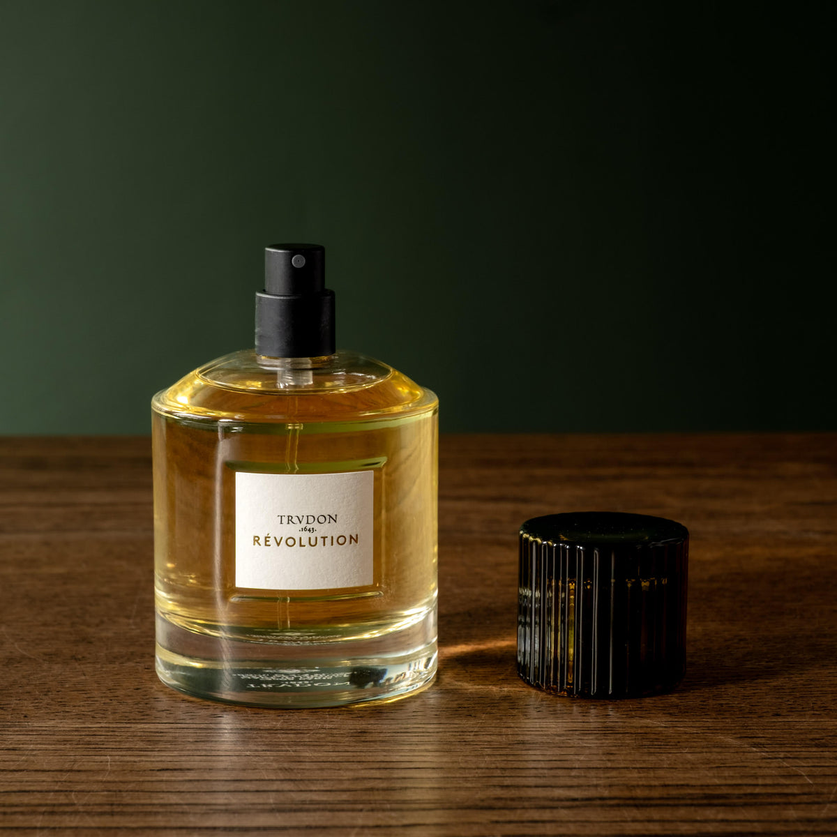 A bottle of Trudon Revolution Eau de Parfum with a black spray top and a separate ribbed black cap, set on a wooden surface against a dark green background.