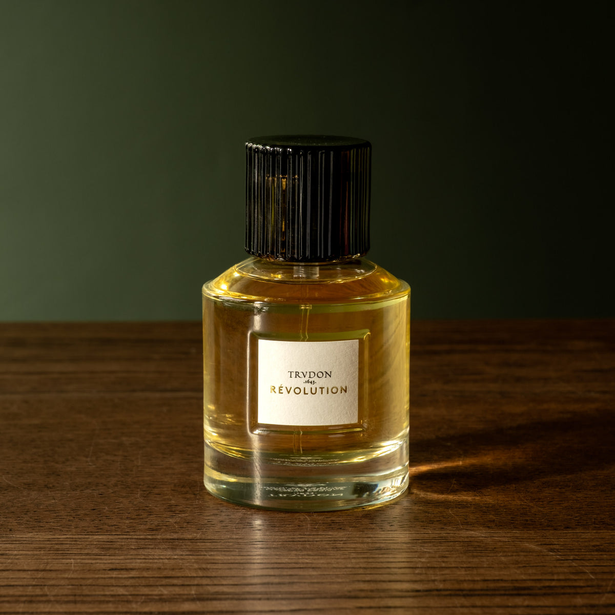 A bottle of Trudon Revolution Eau de Parfum. In a clear glass bottle, a white label and a black ribbed cap, set on a dark wooden surface against a dark green background.
