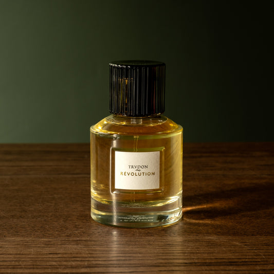 A bottle of Trudon Revolution Eau de Parfum. In a clear glass bottle, a white label and a black ribbed cap, set on a dark wooden surface against a dark green background.