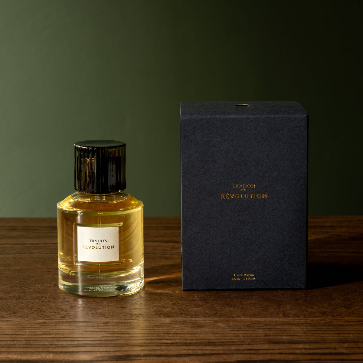 A bottle of Trudon Revolution Eau de Parfum in a glass bottle and a black ribbed cap sits next to its textured navy blue box on a wooden table against a dark green background.