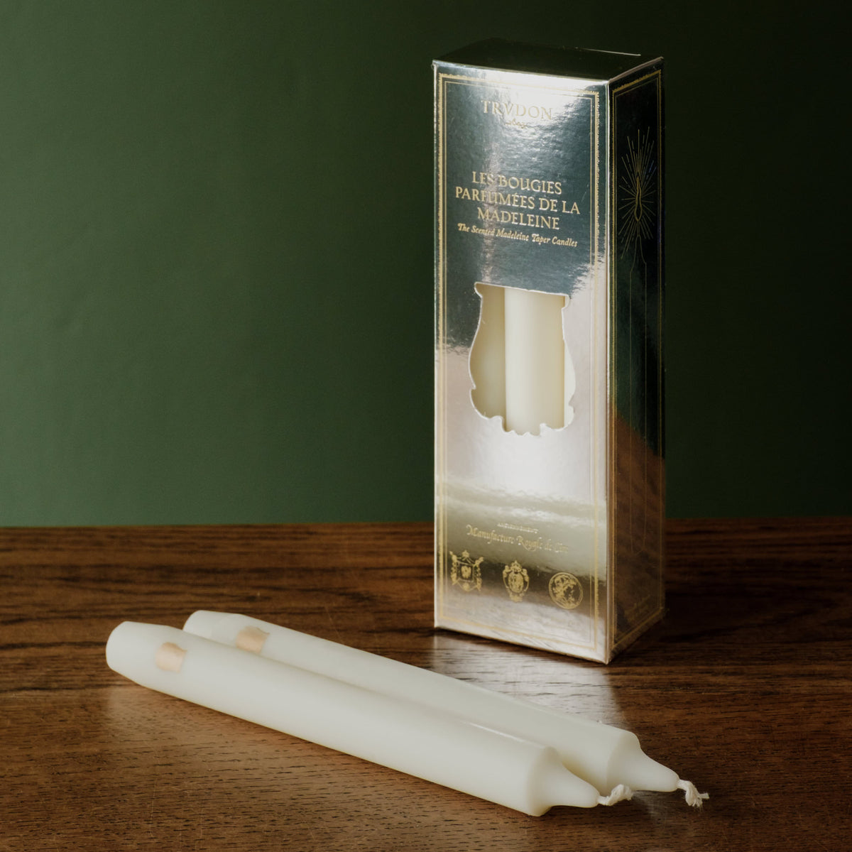 Trudon La Nuit des Glaces Fir scented  6 Taper candles. Showing 2 white taper candles lying on a wooden surface in front of their metallic silver box packaging, set against a dark green background.