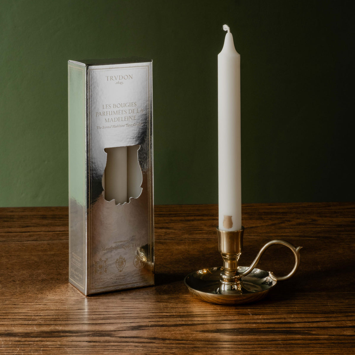 Trudon Fir scented white taper candle in the Trudon brass Dutch Candleholder, sitting beside its branded box of 6 on a wooden table against a dark green wall. 