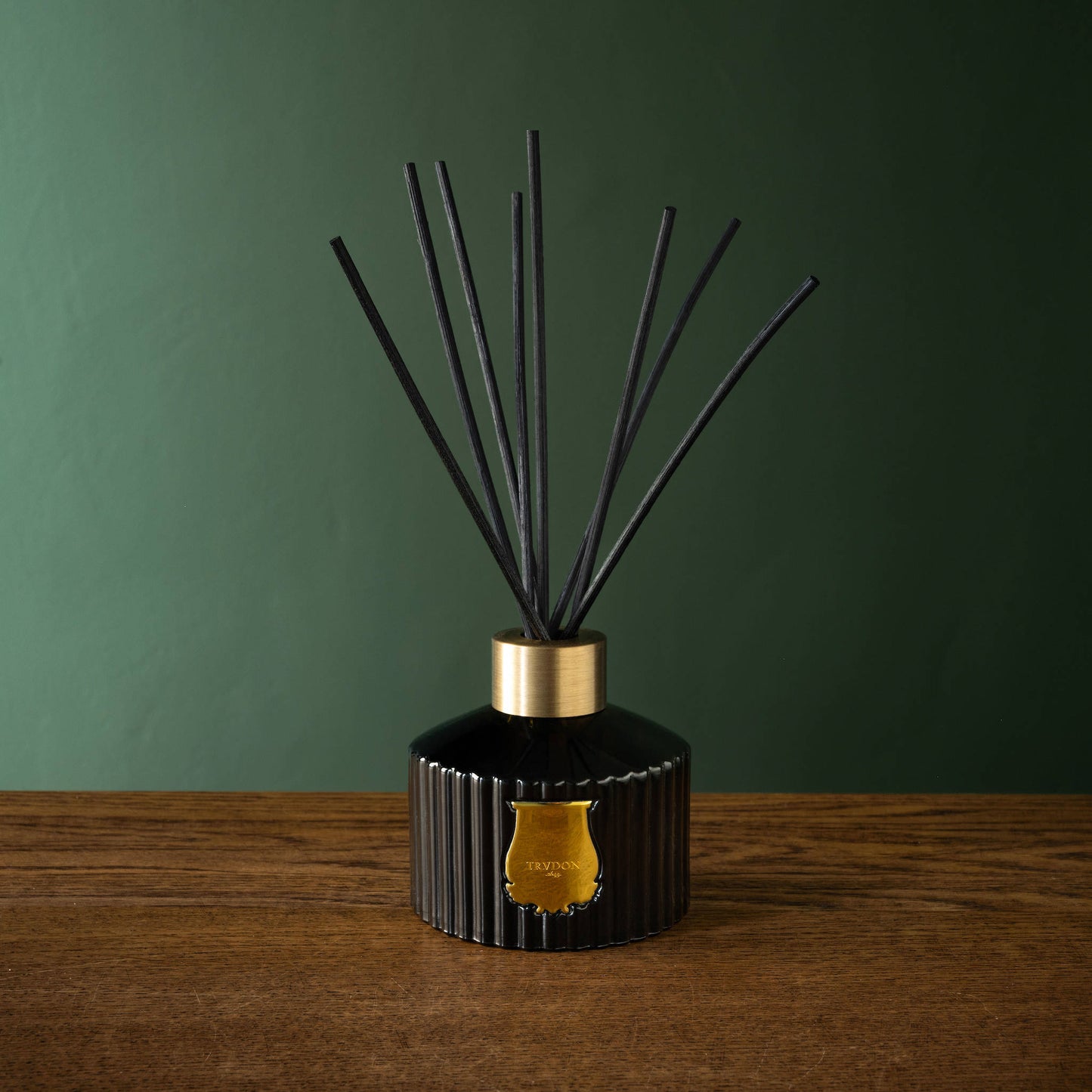Trudon Gabriel Reed Diffuser in dark green glass bottler with gold accents on a wooden surface against a green wall