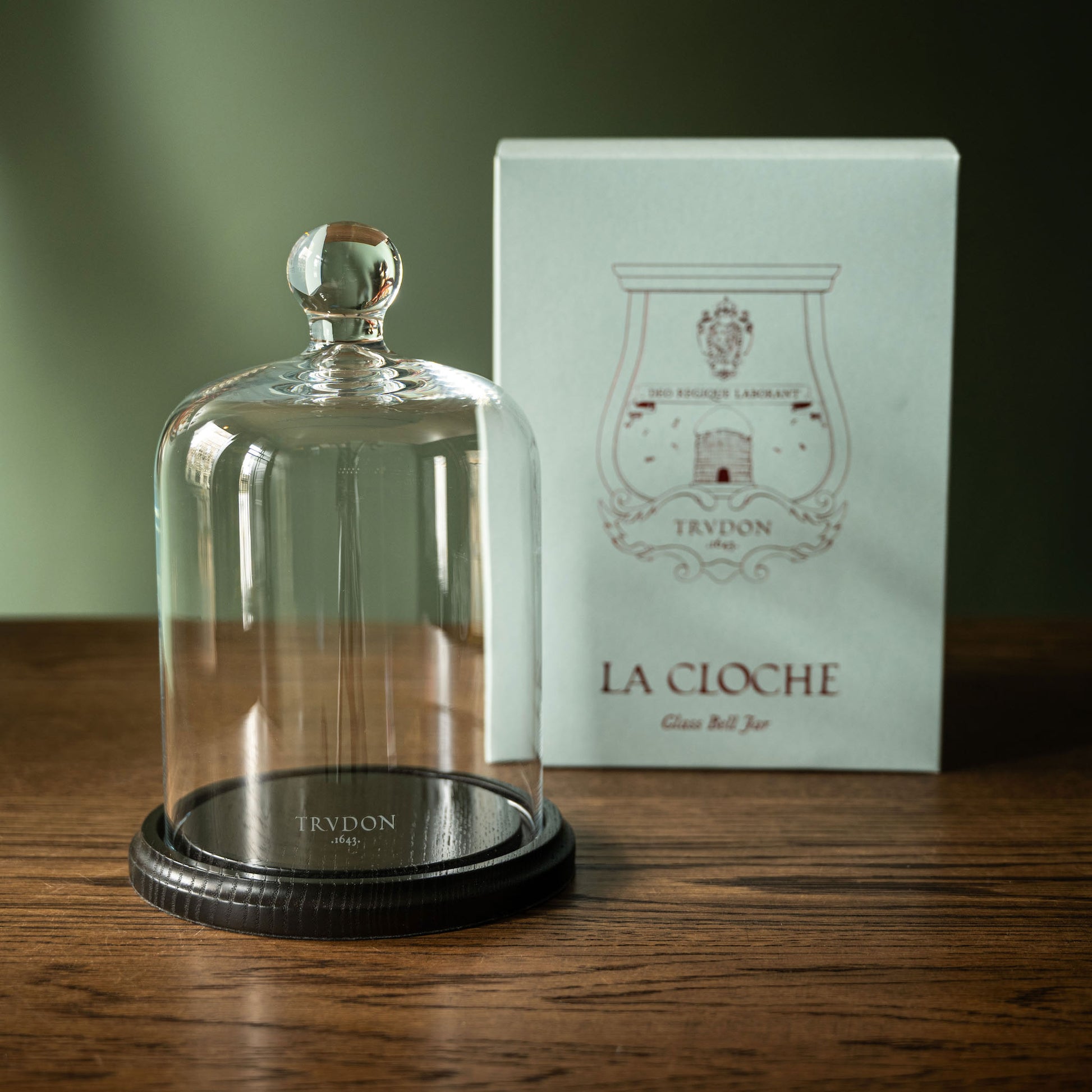 Trudon La Cloche. A glass bell jar with a dark wood base, shown next to it's box on a wooden table.