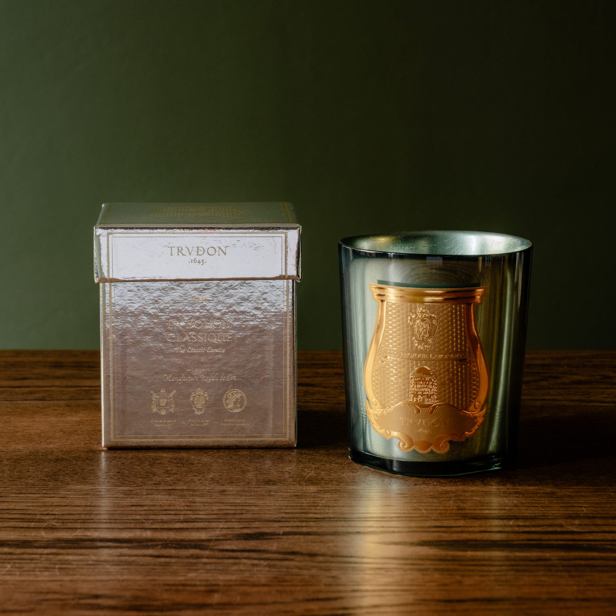 Trudon limited edition La Nuit des Glaces Fir scented 270g candle. Housed in a mirror finish glass jar displayed next to its metallic silver box on a dark wooden table against a dark olive green background.