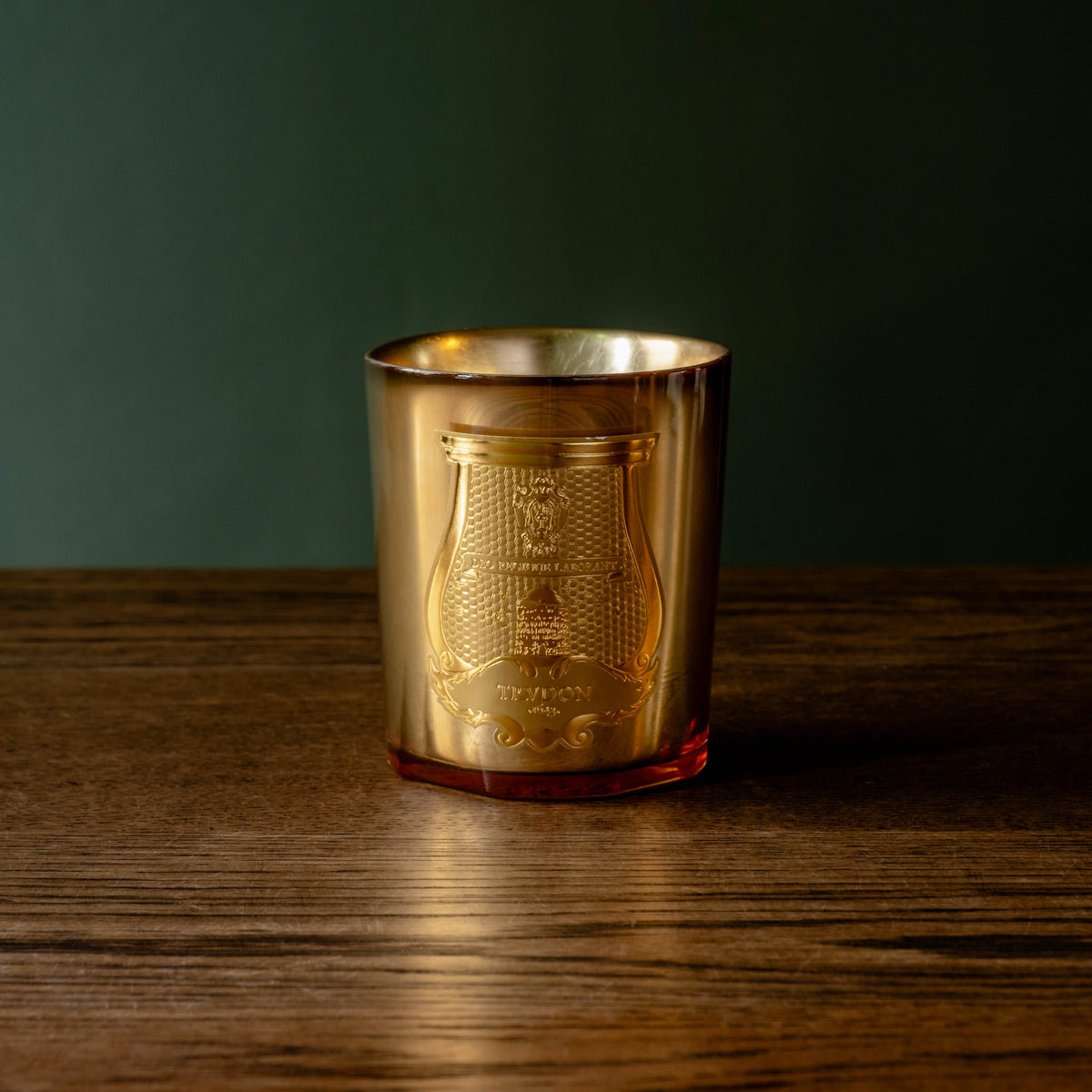 Trudon limited edition La Nuit des Glaces Galerie des Flammes 270g scented candle. Housed in a gold mirror finish glass jar with the Trudon ornate gold crest label. Sitting on a dark wooden table against a dark green background. 