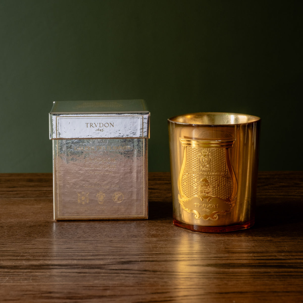 Trudon limited edition La Nuit des Glaces Galerie des Flammes 270g scented candle. Housed in a gold mirror finish glass jar, displayed next to its silver box on a dark wooden table against a dark green background.