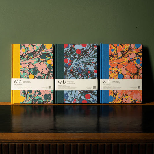 Three Wainwright Bookbinding notebooks with colourful marbled fabric cover designs on a wooden surface against a green wall.